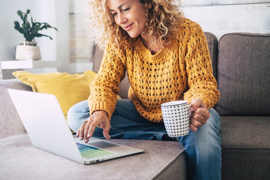 Keltapaitainen nainen istuu sohvalla ja käyttää kannettavaa tietokonetta. Hänellä on kahvikuppi kädessään.