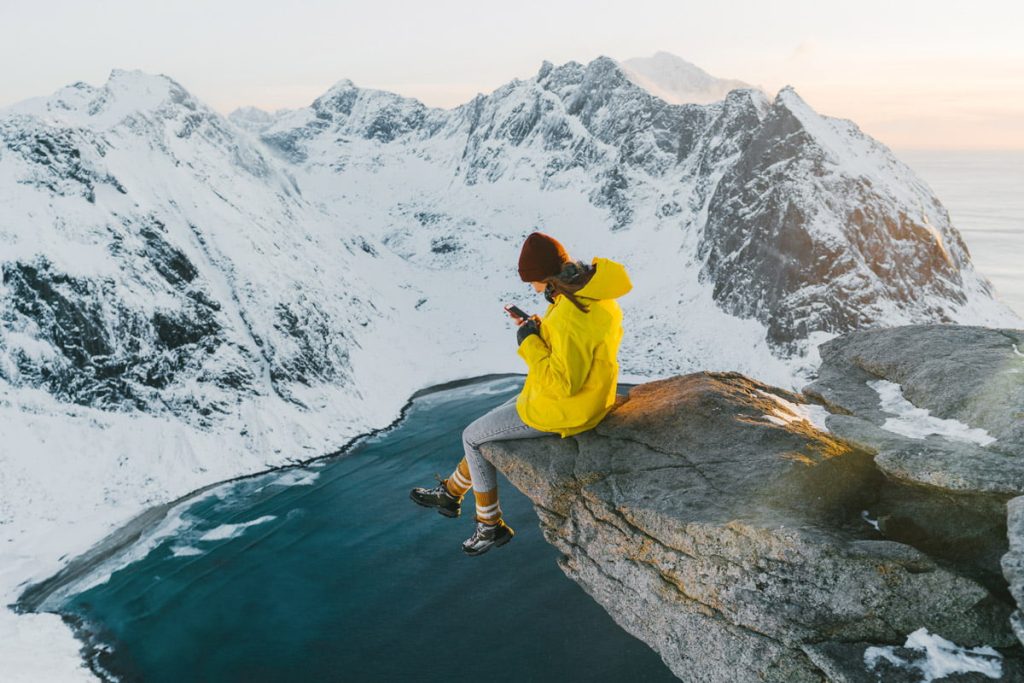 Talvinen vuoristomaisema, jossa vuoren rinteellä istuu keltatakkinen nainen puhelinta katsoen.