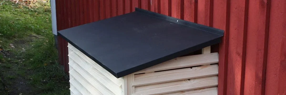 Ilmalämpöpumpun suojakotelo, ilmalämpöpumpun ulkoyksikön puun värinen suojakotelo punaisen puutalon seinällä. Suojakotelossa on musta metallinen kansi.