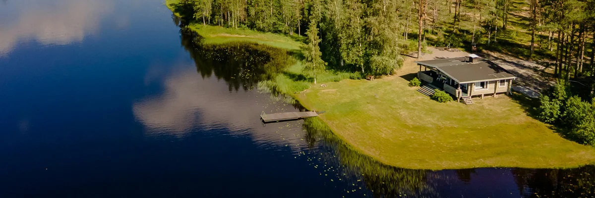 Vehreä mökkitontti ylhäältä kuvattuna. Yksikerroksinen mökki aivan sinisen ja tyynen järven rannalla.